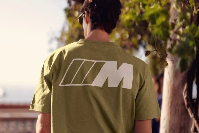 Man wearing an olive green T-shirt with a large white BMW M logo on the back, standing outdoors near a tree.