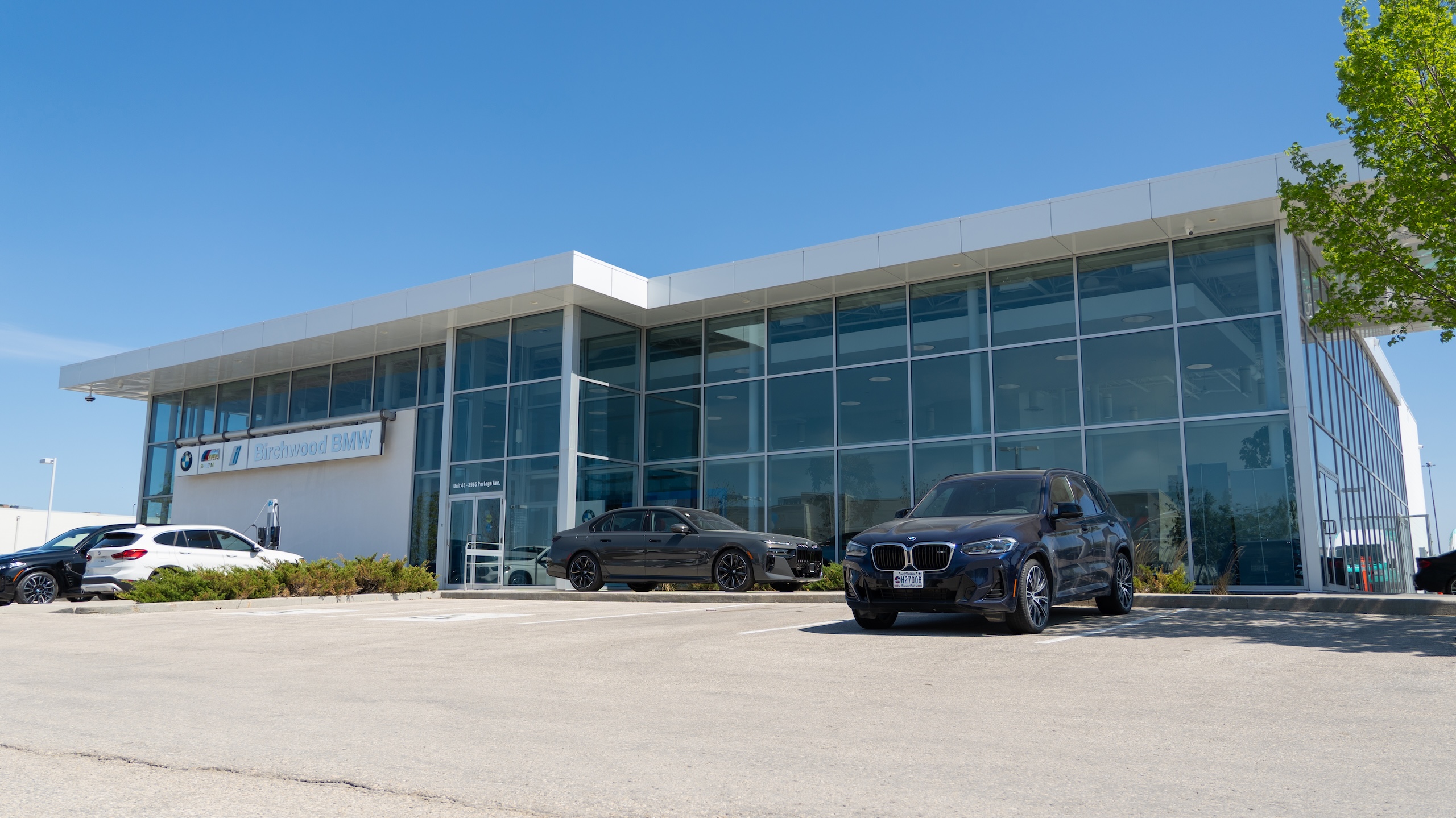 Exterior of Birchwood BMW dealership under a clear blue sky, with modern glass-front architecture and several BMW vehicles parked in front, including a white SUV and a black sedan.
